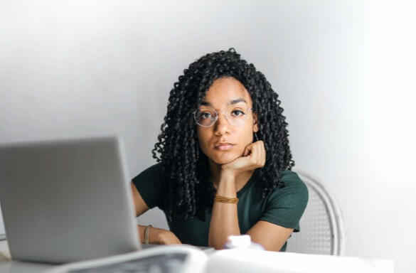 Young brown skinned woman with long spirally hair sat at laptop looking despondent